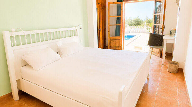 Helles Schlafzimmer mit weißem Doppelbett, Terrakottaboden und Terrassentür mit Poolblick im Agroturismo Ses Salines