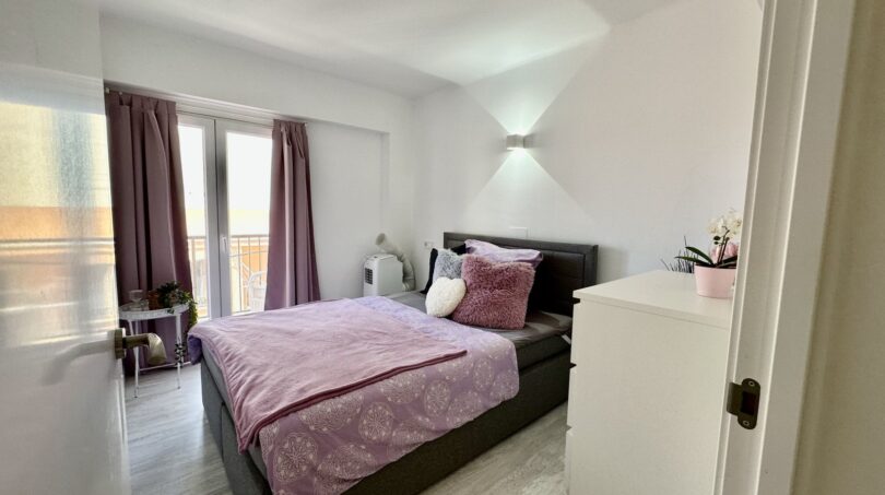 Helles Schlafzimmer mit Doppelbett, lila Bettwäsche, Wandleuchten und Balkonzugang mit Ausblick