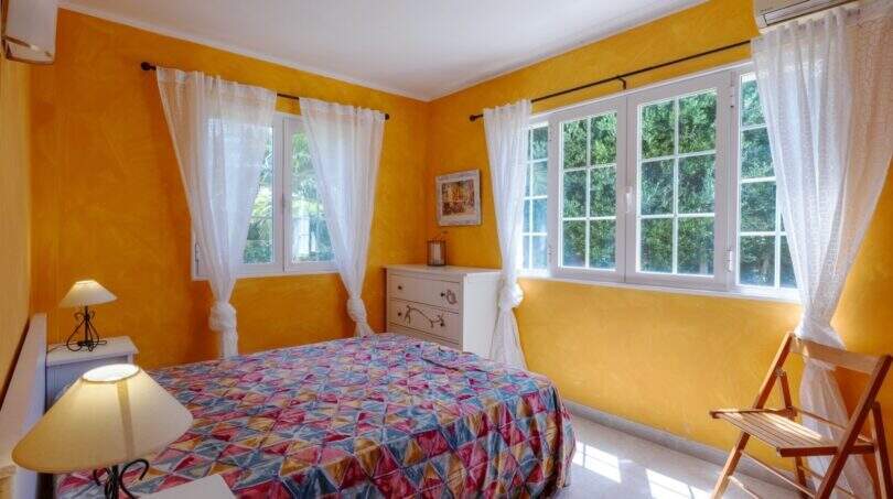 Helles Schlafzimmer mit gelbem Wandanstrich, bunter Tagesdecke und zwei Fenstern mit Gardinenschmuck