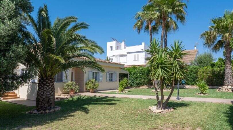 Außenansicht mediterraner Bungalow in Llucmajor mit gepflegtem Garten, Palmen und weißer Villa im Hintergrund.