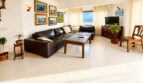 Helles Wohnzimmer des Penthouses mit Ledersofa, Gemälden, Pflanzen und Meerblick durch große Fenster.