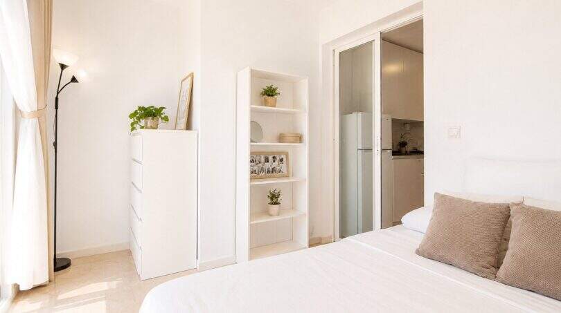 Helles Schlafzimmer im Penthouse Palma de Mallorca mit weißem Bett, Regal und Blick in offene Küche.