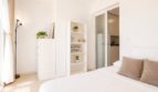 Helles Schlafzimmer des Penthouses in Palma mit weißem Bett, Regal und Blick in offene Küche.