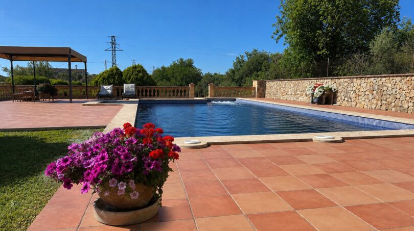 Finca Sencelles: Gepflasterter Poolbereich mit Blumentopf, Steinmauer und Pergola unter blauem Himmel