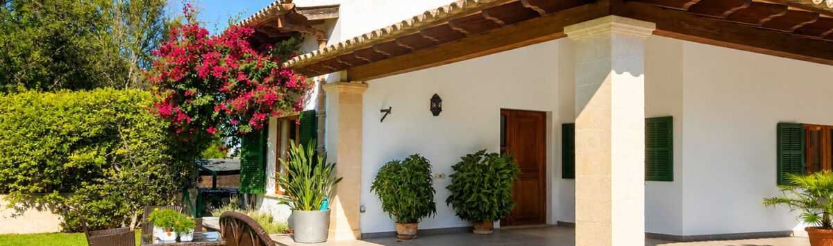 Außenansicht der Finca Sencelles: weißes Landhaus mit Terrakottadach, Säulenveranda, gepflegtem Rasen und bunten Blumen.
