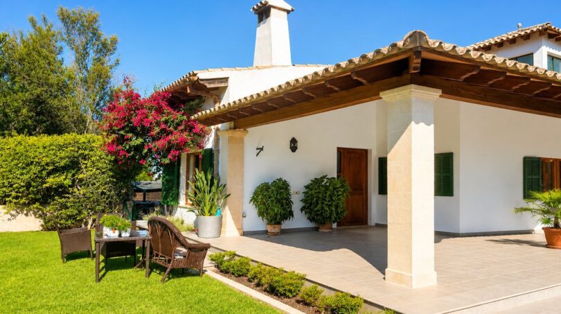 Außenansicht der Finca Sencelles: weißes Landhaus mit Terrakottadach, Säulenveranda, gepflegtem Rasen und bunten Blumen.