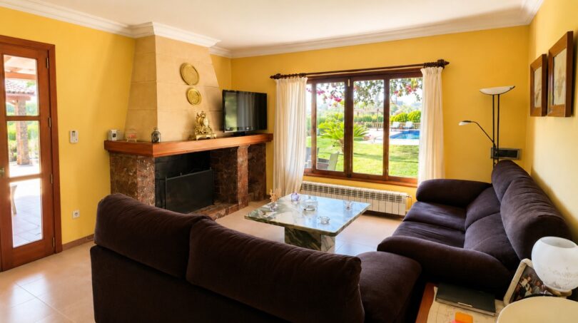 Wohnzimmer der Finca mit Kamin, dunklen Sofas und Blick auf Pool und Garten