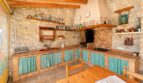 Rustikale Küche der Finca mit Steinwand, Holzregalen voller Flaschen, Kamin und türkisen Vorhängen.