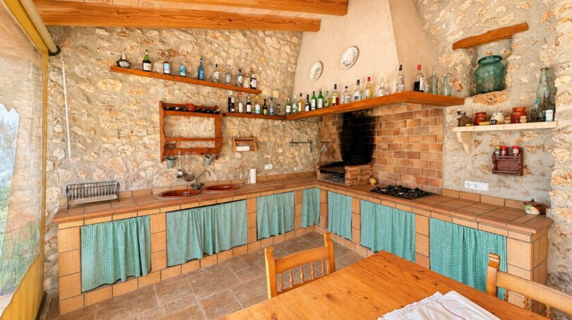 Rustikale Küche der Finca mit Steinwand, Holzregalen voller Flaschen, Kamin und türkisen Vorhängen.
