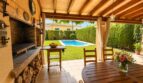 Überdachte Terrasse mit Grillkamin und Holztisch, Blick auf gepflegten Garten mit Pool der Doppelhaushälfte in Llucmajor