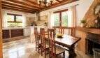 Rustic dining room of the Llucmajor finca with wooden beam ceiling, stone wall, fireplace and adjacent kitchen area