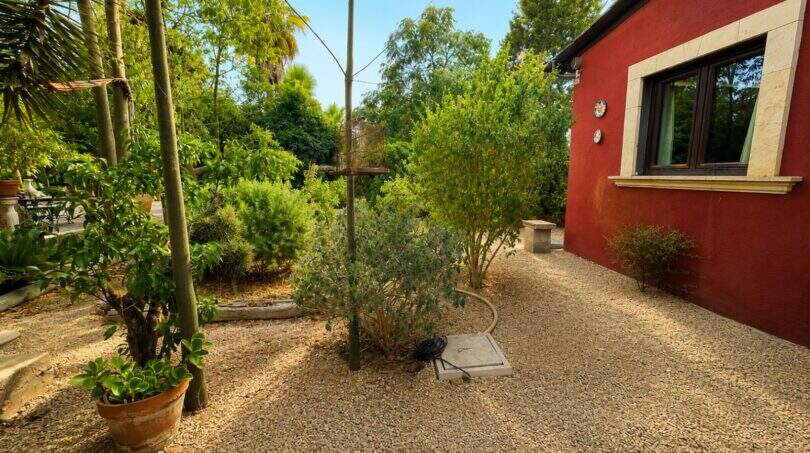 Gravel path next to red countryside house with natural stone frame window, lush garden and Mediterranean plants, Finca Llucmajor