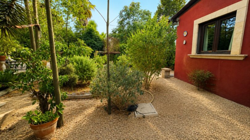 Kiesweg neben rotem Landhaus mit Natursteinrahmen-Fenster, umgeben von mediterranem Garten mit Palmen und Sträuchern