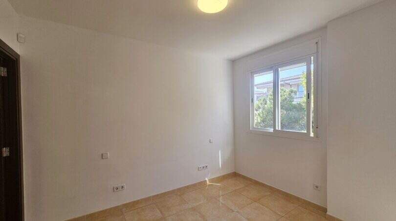 Helles, leeres Schlafzimmer der Wohnung in Porto Cristo mit Fliesenboden, weißen Wänden und Fenster zum Nachbargebäude.