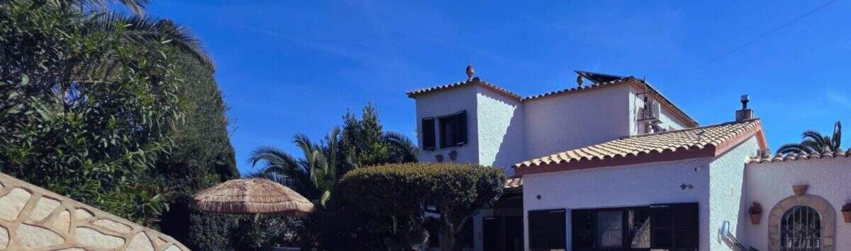 Außenansicht: Mediterranes Ferienhaus in Cala Romántica mit blauem Pool, Sonnenterrasse und üppigem Grün unter strahlend blauem Himmel.