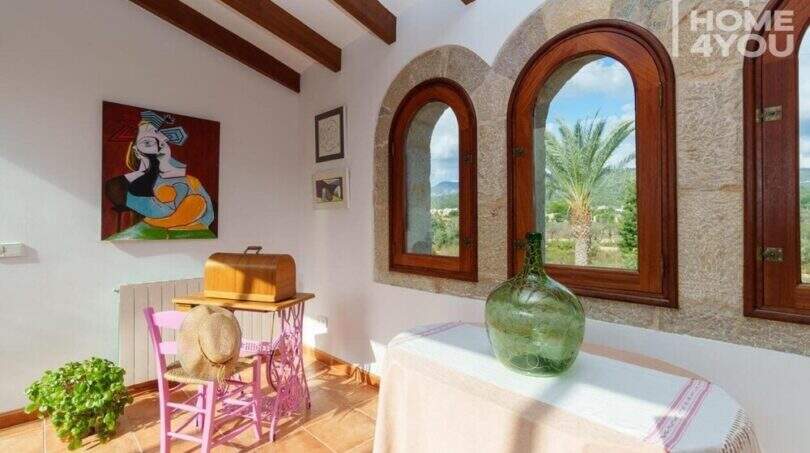 Wohnzimmer der mallorquinischen Villa mit Natursteinbögen, Holzbalken und Blick auf Palmen und Tramuntana-Gebirge.