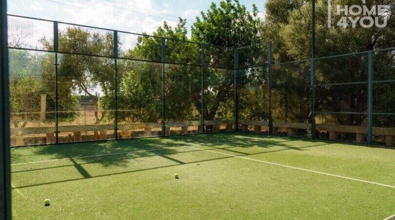 Pista de pádel de la finca con césped artificial, paredes de cristal y red de metal, rodeada de vegetación mediterránea.