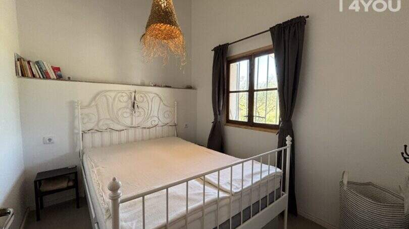 Bedroom of Finca Felanitx with white metal bed, rattan lamp, bookshelf and window with dark curtains.