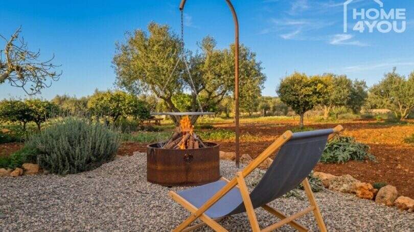 Garden area of Finca Felanitx with fireplace, sun lounger and olive trees under blue sky.