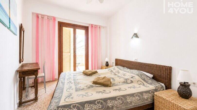 Luminoso dormitorio con cama doble, cabecero de ratán, cortinas rosas, acceso al balcón y ventilador de techo en Ses Salines