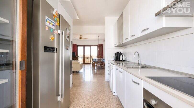 Cocina moderna con refrigerador de acero inoxidable y armarios empotrados blancos, vista a la luminosa sala de estar con acceso a la terraza