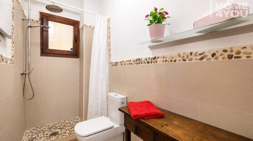 Luminoso baño con ducha, suelo de guijarros, estantería de madera y planta decorativa en apartamento en Ses Salines.