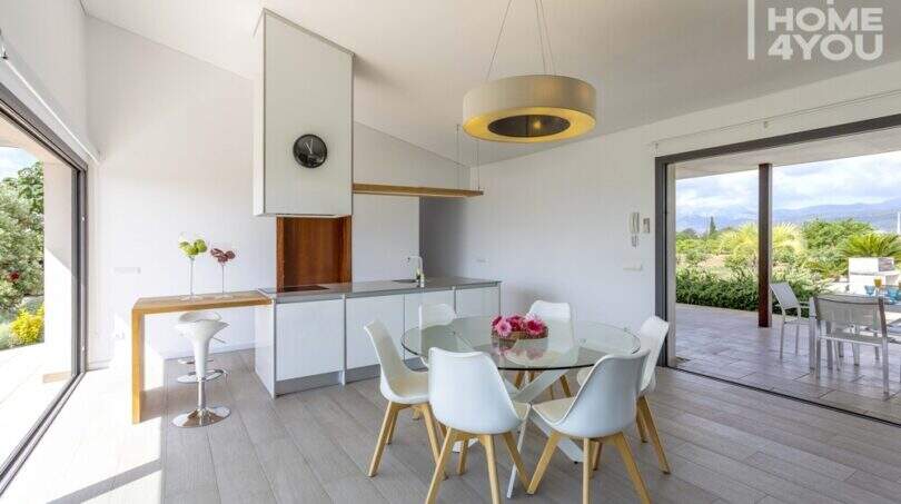 Comedor luminoso con mesa redonda de cristal, sillas blancas y cocina abierta, vista al jardín mediterráneo