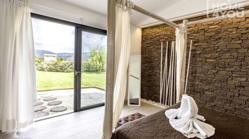 Dormitorio con cama con dosel, pared de piedra natural y acceso a terraza al jardín cuidado de la finca mediterránea.