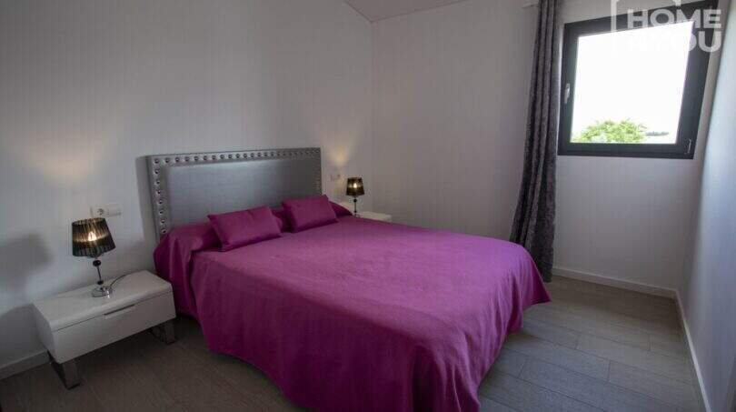 Dormitorio de la finca en Sa Pobla con cama doble, ropa de cama morada, cabecero plateado y ventana con cortinas.