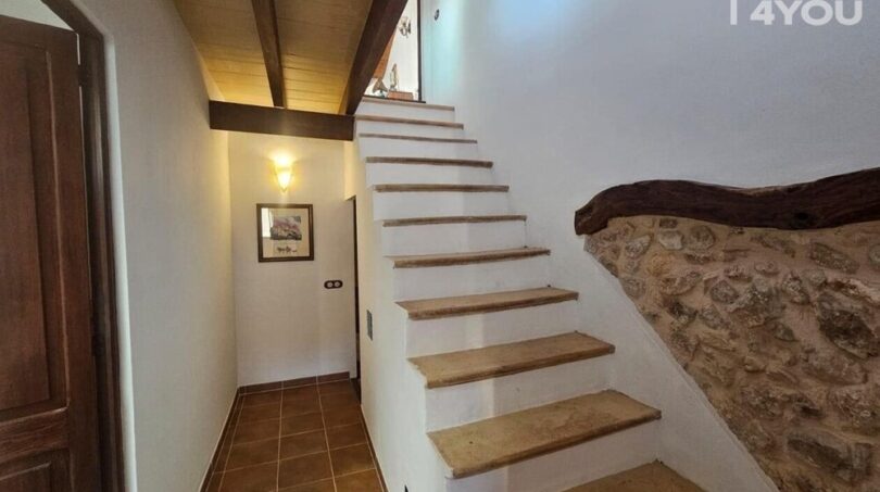 Escalera rústica con peldaños de madera, pared de piedra natural y techo de vigas de madera en casa de pueblo Petra