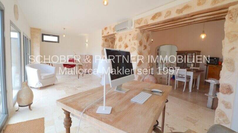 Helles Wohnzimmer mit Natursteinwand, Holzbalken und modernem Arbeitsbereich im sanierten Landhaus Portocristo.