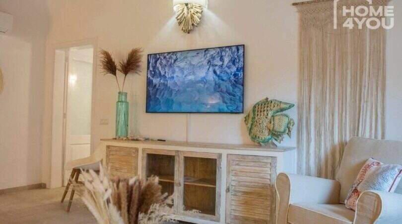 Helles Wohnzimmer mit TV, Holzkommode, Sessel & Boho-Deko im Bungalow in Alcúdia, Mallorca.