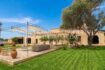 Außenansicht Landhaus Mallorca: Rustikale Natursteinfassade mit gepflegtem Rasen, Sonnensegel-Terrasse und altem Olivenbaum