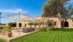 Außenansicht Landhaus Mallorca: Rustikale Natursteinfassade mit gepflegtem Rasen, Sonnensegel-Terrasse und altem Olivenbaum