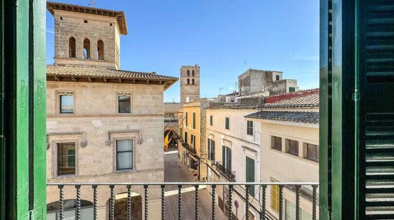 Balcón con contraventanas verdes de madera y vistas a fachadas históricas de piedra y campanarios de iglesia en Felanitx