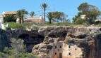 Historische Felshöhlenbehausung in Cala Murada mit modernen Villen und Palmen im Hintergrund.