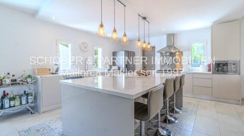 Moderne weiße Einbauküche mit großer Kochinsel, Barhockern und Hängeleuchten in mediterraner Villa Sa Coma