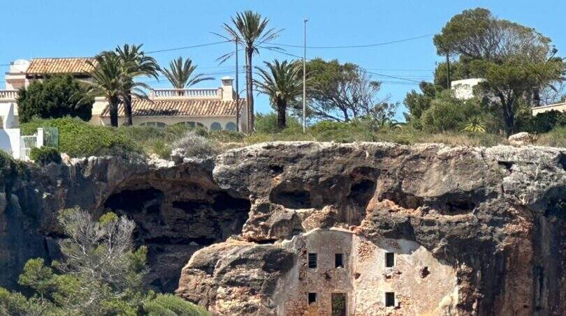 Historische Felshöhlen-Ruine in Cala Murada mit mediterraner Villa und Palmen im Hintergrund.