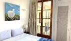 Dormitorio luminoso con cama doble, colcha Ikat, cuadro de ovejas y balcón de madera con vistas al casco antiguo de Palma.