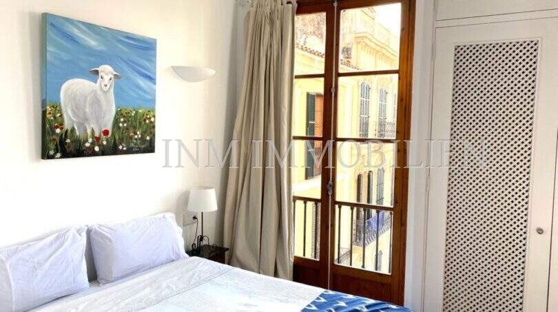 Dormitorio luminoso con cama doble, colcha Ikat, cuadro de ovejas y balcón de madera con vistas al casco antiguo de Palma.