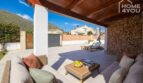 Covered terrace with lounge furniture, natural stone wall and view of sun terrace with sun loungers and mountain panorama.