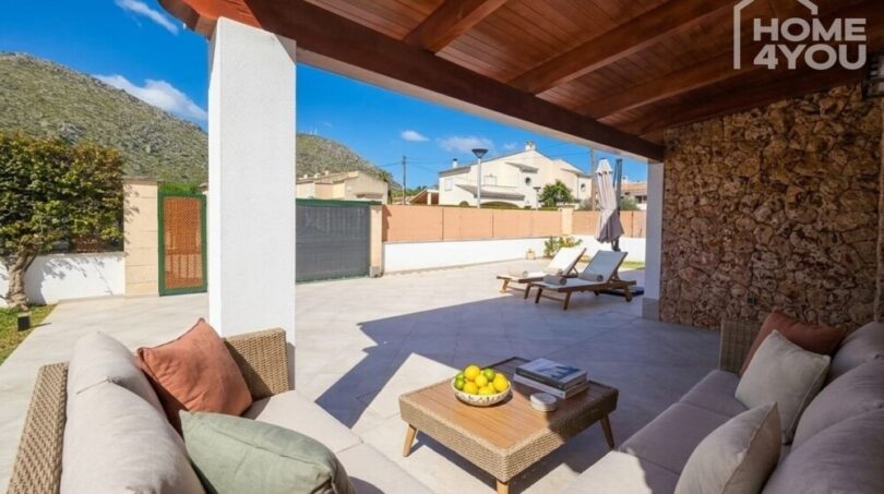 Covered terrace with lounge furniture, natural stone wall and view of sun terrace with sun loungers and mountain panorama.