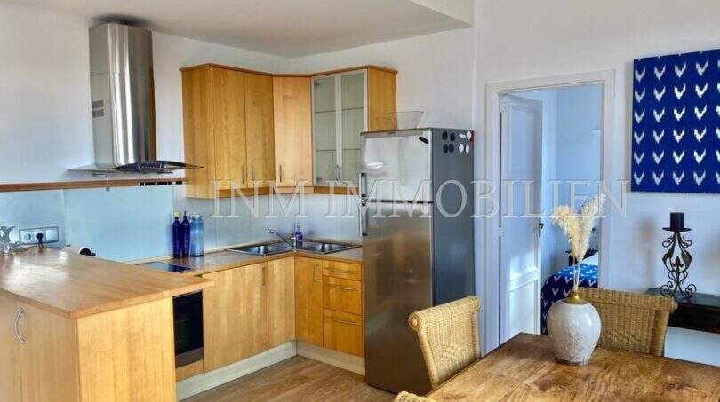 Cocina abierta con frentes de madera y frigorífico de acero inoxidable, zona de comedor adyacente con sillas de ratán en apartamento del casco antiguo de Palma