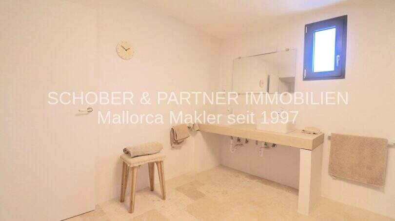 Helles Badezimmer mit cremefarbenem Natursteinboden, modernem Waschtisch und kleinem Holzhocker im sanierten Landhaus Portocristo
