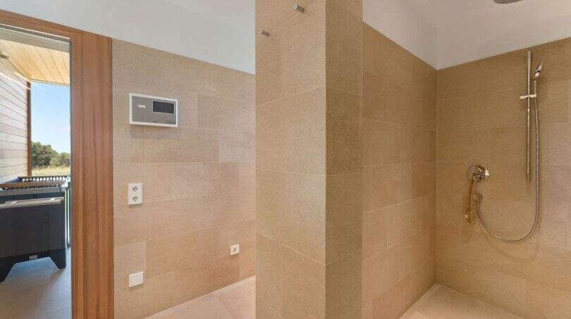 Modern bathroom with beige natural stone tiles, accessible shower with rain shower head and view of the terrace.
