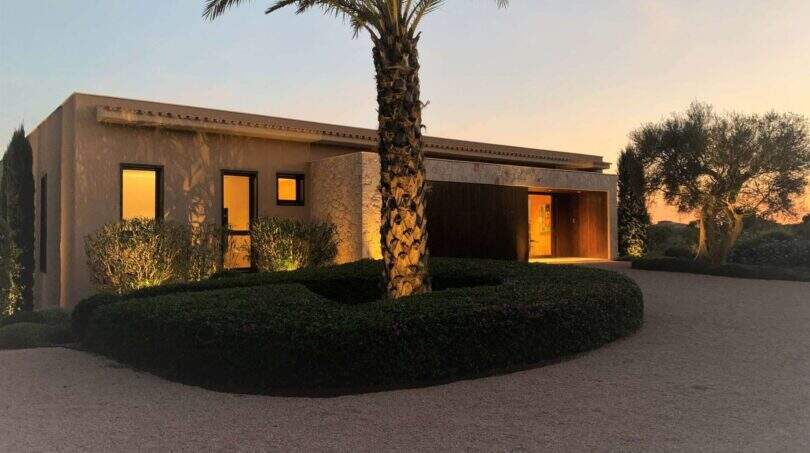 Exterior view design house S'Alqueria Blanca at sunset with palm, circular path and warm light ambiance.