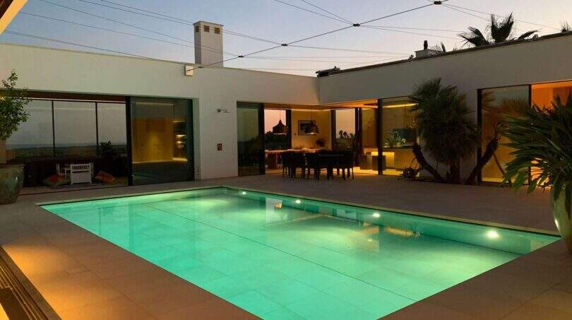 Illuminated outdoor pool at dusk, surrounded by modern white facade with panoramic glazing in S'Alqueria Blanca.