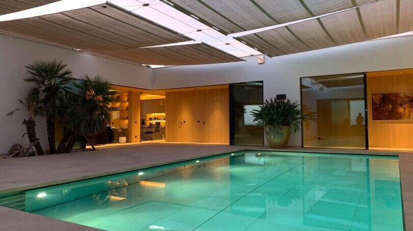 Indoor pool area with turquoise water, palms and modern living access in designer villa S'Alqueria Blanca
