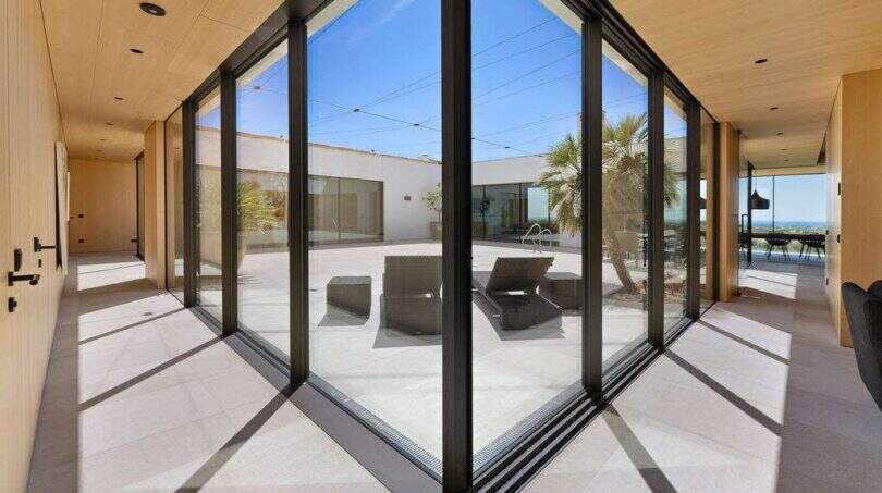 Light-filled glass corridor with view of courtyard terrace with lounge chairs and sea view in modern design house