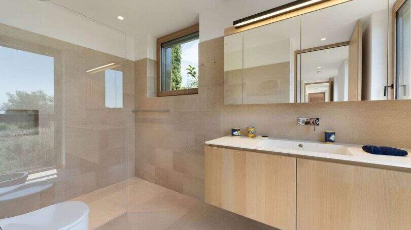 Modern bathroom with light wooden vanity, natural stone tiles and large mirror in design house S'Alqueria Blanca